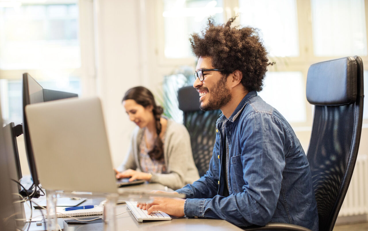 Lächelnder Mitarbeiter am Computerarbeitsplatz, im Hintergrund eine Kollegin